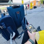 Blue suitcases sitting on a sidewalk, with a novel and a yellow folding fan in the foreground.
