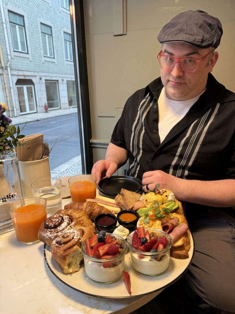 Fes, sitting in a cafe, looking dubiously at the camera, while an enormous tray of cafe foods sits before him.